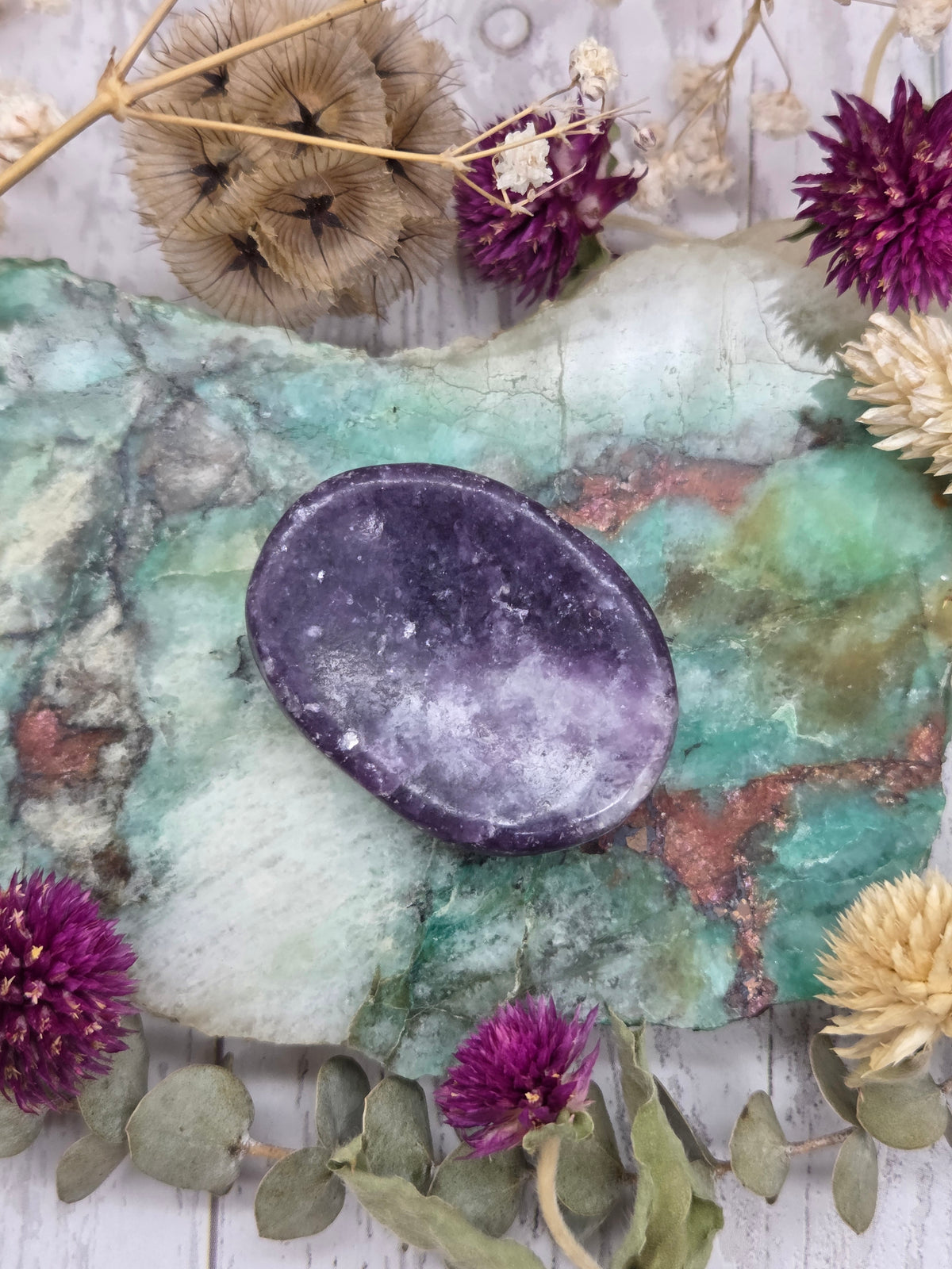 A polished purple Lepidolite worry stone on a colorful background with dried flowers.