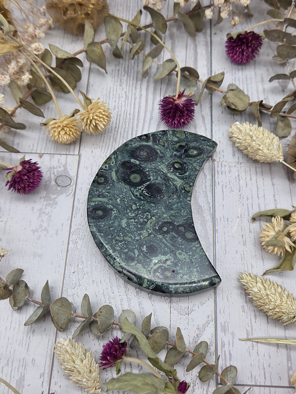 Kambaba Jasper moon carving on a wooden surface surrounded by dried flowers